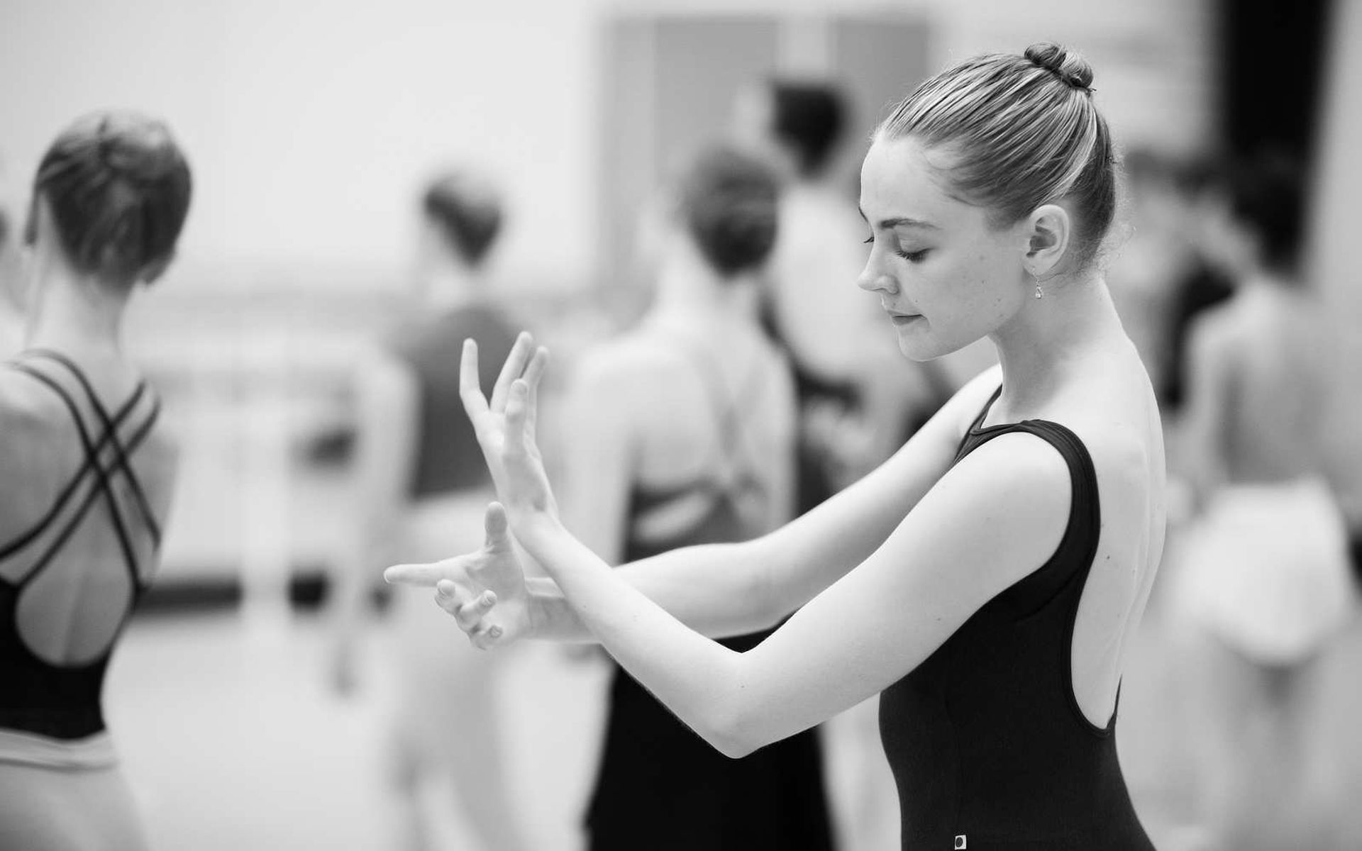 En balletdanser i sort trikot står i profil og øver hænder og arme. Den ene arm er bøjet foran kroppen, mens den anden hånd former eller strækker fingrene. Udtrykket virker koncentreret og roligt. Bagved ses flere dansere i træningstøj, let ude af fokus, som i en øvesal. Billedet er i sort-hvid og skaber en blød, rolig stemning, hvor kroppens linjer fremhæves.