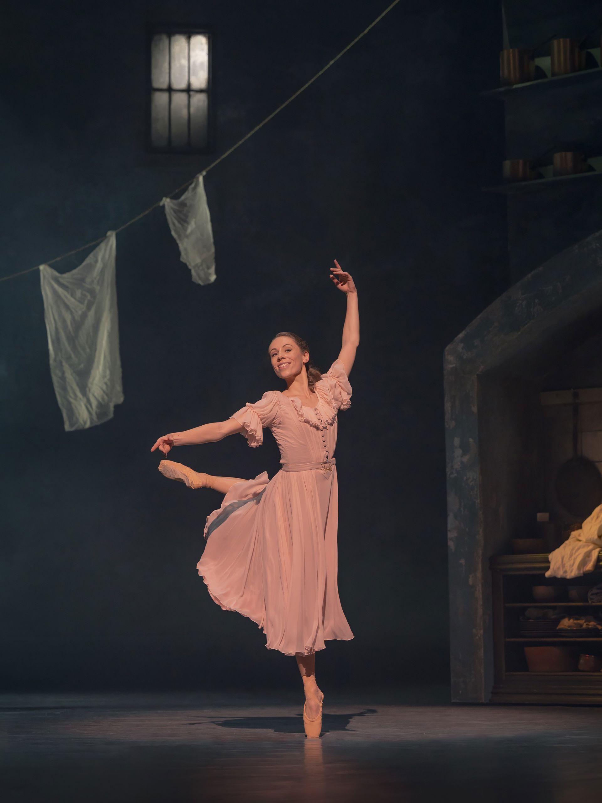 En balletdanser i lys rosa kjole danser alene på en mørk scene. Den ene arm er løftet højt, og kroppen er let drejet i en elegant positur. I baggrunden hænger lyse stykker vasketøj på snore, som giver scenen et enkelt og poetisk udtryk.