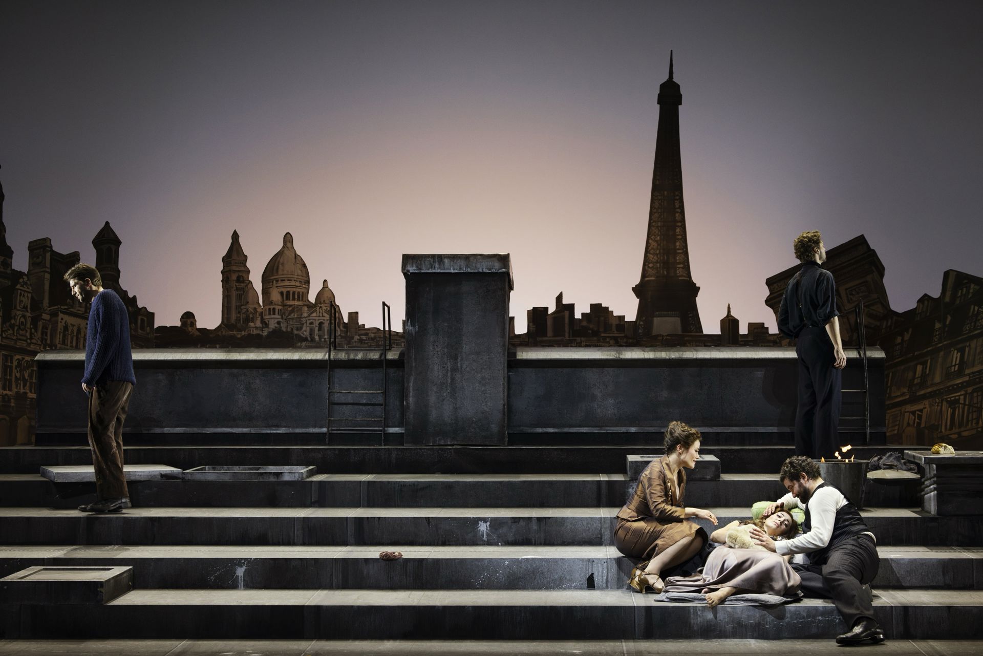 To personer sidder på trapperne foran en kulisse med Paris' skyline og Eiffeltårnet i baggrunden, hvor den ene ligger ned med støtte fra den anden. Til venstre står en person med ryggen til, og til højre står en anden person opret ved siden af en lille ildtønde, mens himmelen toner i dæmpede skumringsfarver.