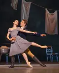 To balletdansere danser tæt sammen på en scene med vasketøj hængende i baggrunden. Den ene danser i gråblå kjole løftes eller støttes af den anden, som står bagved med armen omkring kroppen. Scenen har varmt lys og et hverdagsligt, romantisk udtryk.