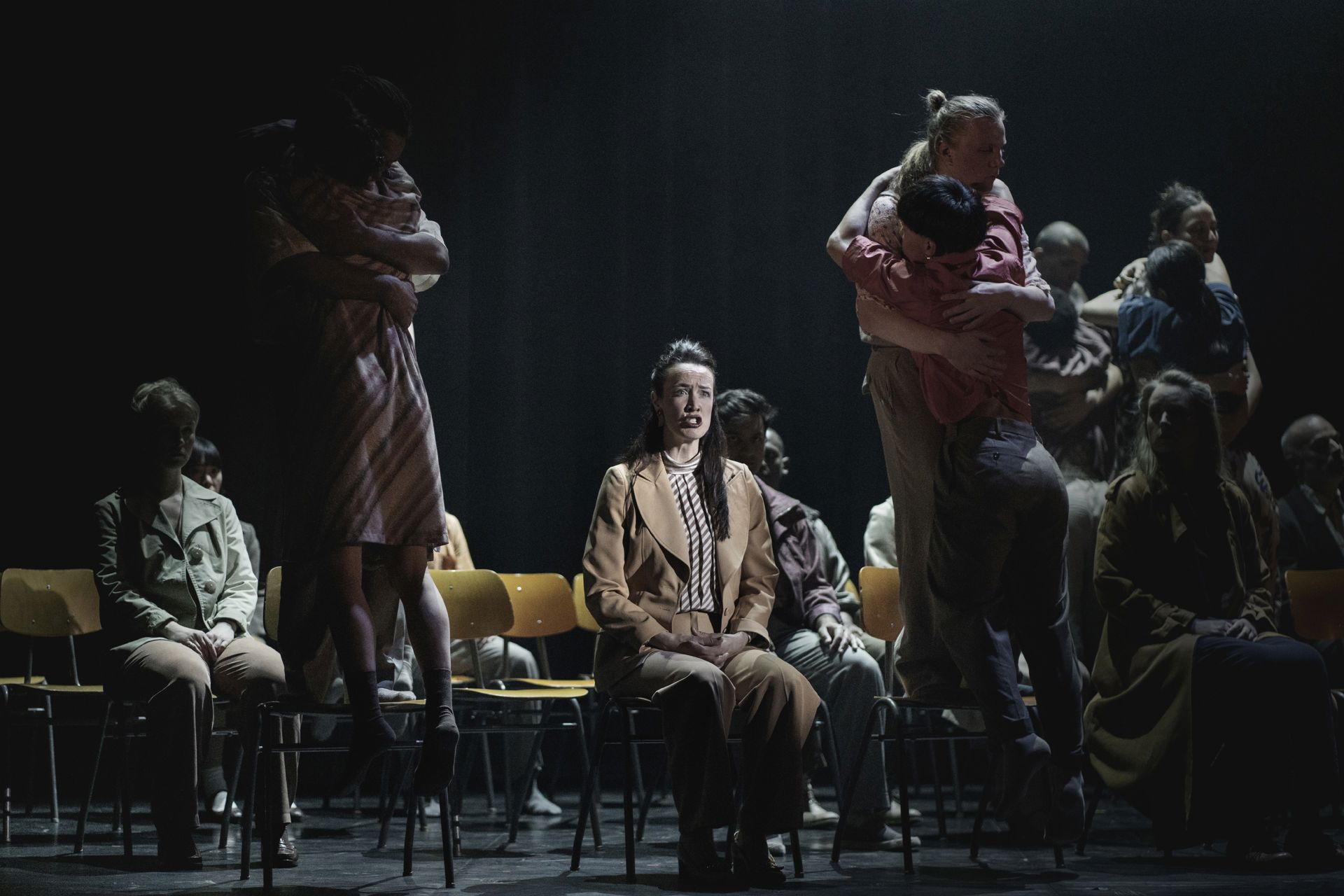 En teaterscene med en gruppe mennesker, der sidder og står på rækker af stole i dunkelt lys.  Midt i billedet sidder en person oplyst af en spot med hænderne i skødet.  Omkring står og sidder andre tæt sammen, nogle holder om hinanden. Stemningen er alvorlig og melankolsk.