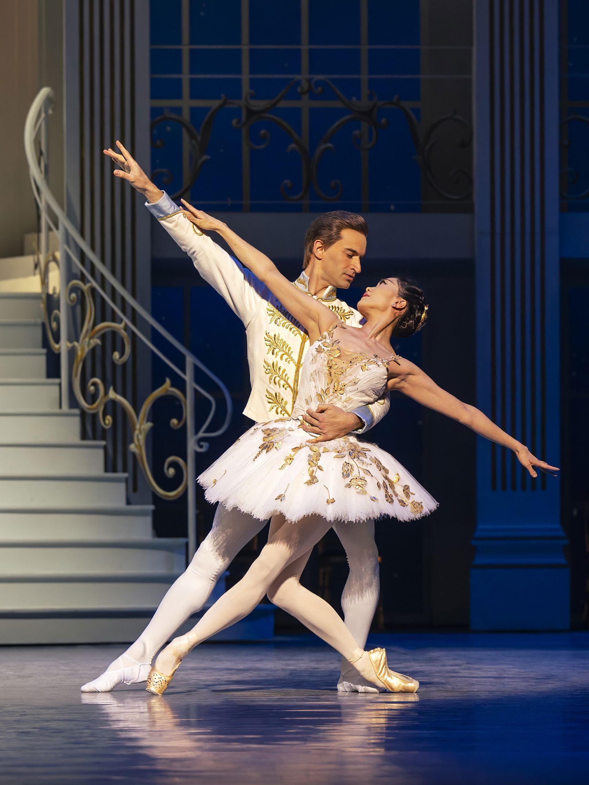 To balletdansere i lyse kostumer danser tæt sammen foran en trappe med blå vinduer i baggrunden. Den ene støttes i en skrå, strakt position med armen ud til siden, mens den anden holder om kroppen. Scenen har et klassisk og romantisk balletudtryk.
