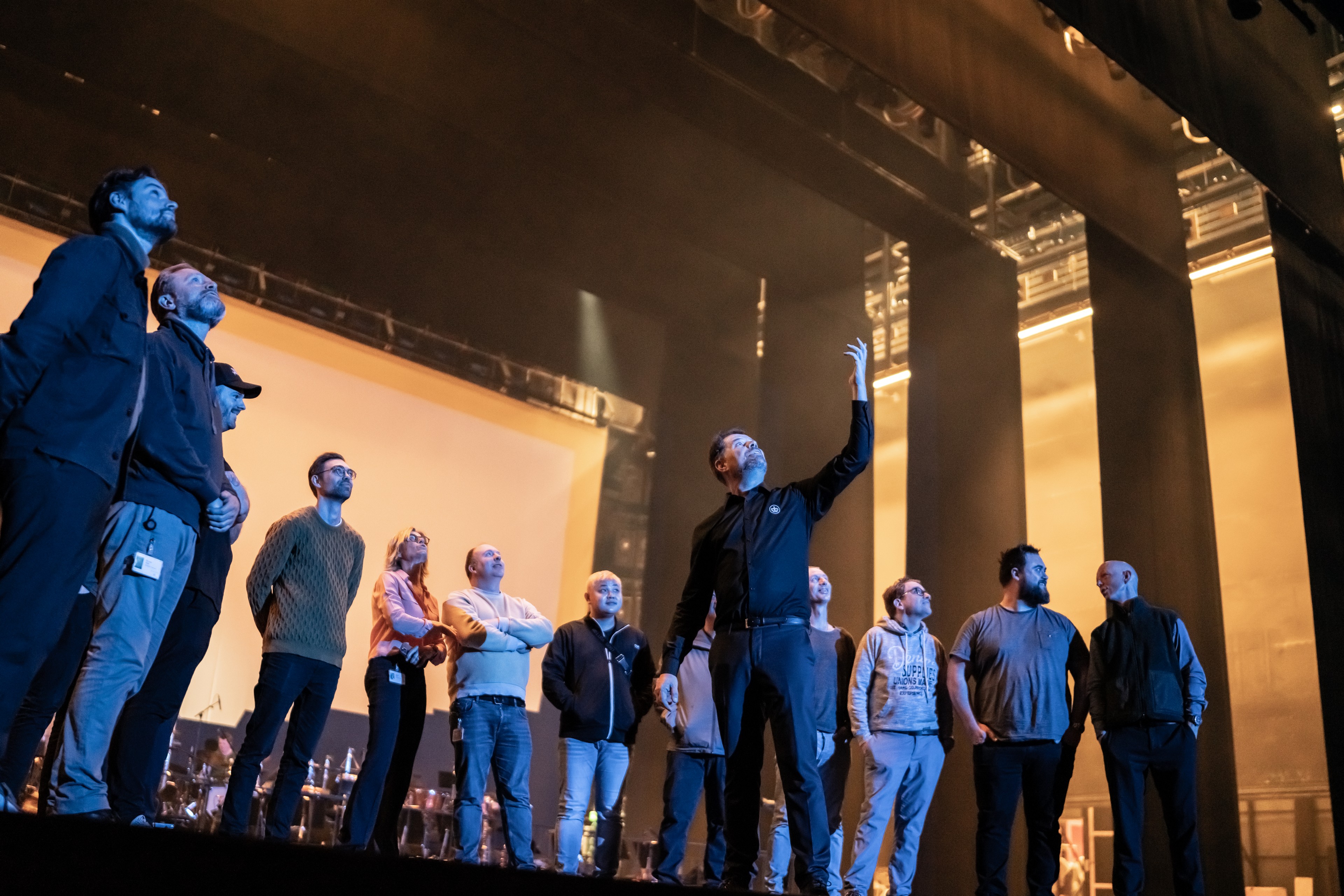 En gruppe mennesker står på store scene i Operaen. Scenen er oplyst med varmt, gyldent lys fra siden og bagfra, og der ses høje, mørke scenestrukturer og lysrigge over dem. Gruppen står på række i almindeligt hverdagstøj. Midt i gruppen har en person den ene arm løftet, som om noget forklares eller peges ud. De øvrige ser i samme retning. I baggrunden anes musikinstrumenter og udstyr.