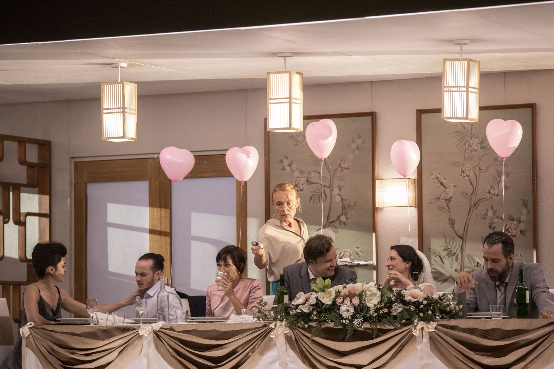 En teaterscene med flere personer samlet ved et langt festbord med blomster, glas og lyserøde hjerte balloner. Personerne sidder tæt sammen og ser ud til at være midt i en samtale. De lyse vægge, papirlamper og blomstrede dekorationer giver scenen et bryllups- eller selskabspræg.