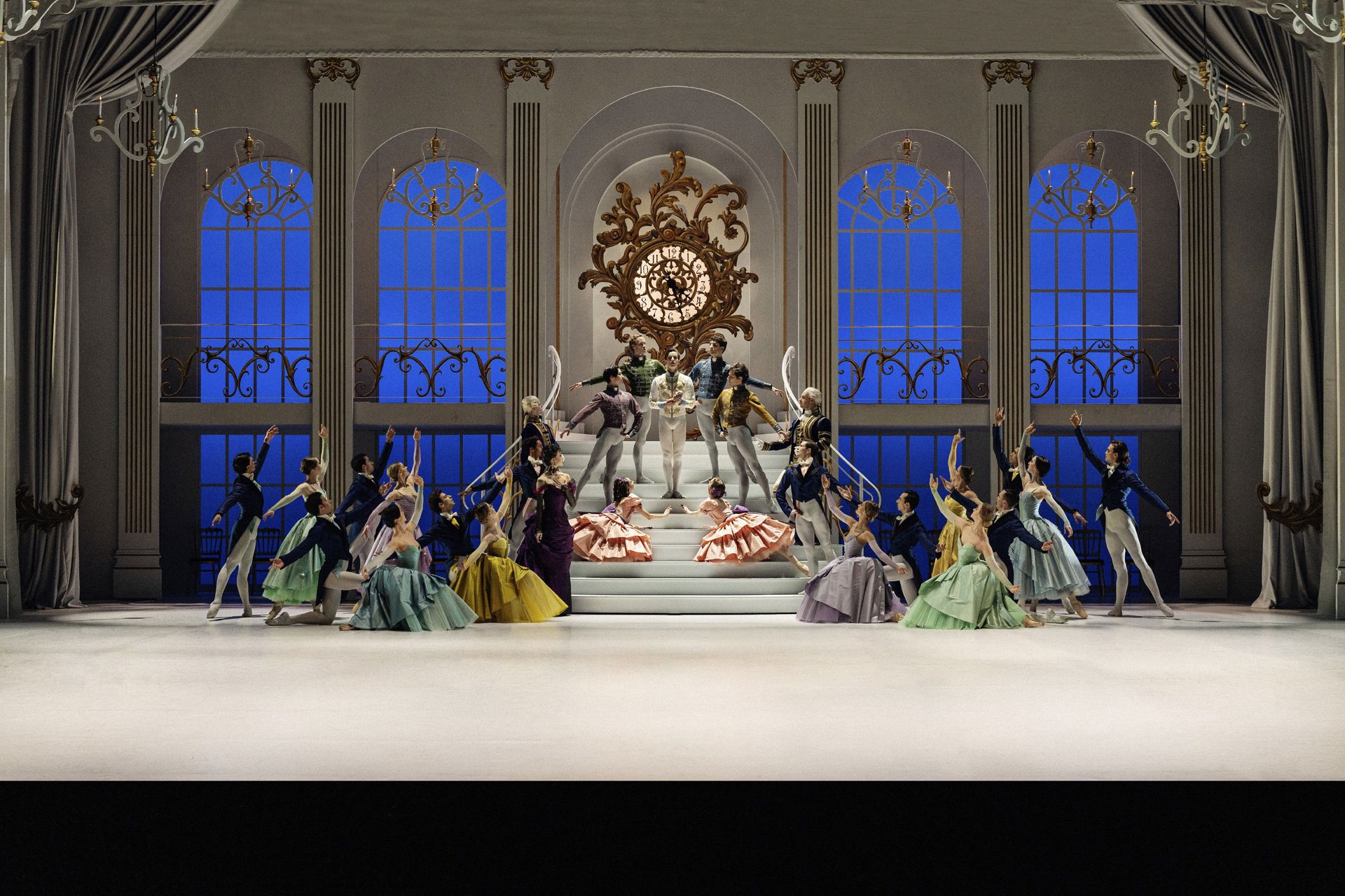 En stor balletscene med mange dansere i farverige historiske kostumer. Scenografien viser en lys sal med høje blå vinduer, trapper og et stort dekorativt ur i midten. Danserne er fordelt i grupper omkring scenen med løftede arme og klassiske balletpositurer.