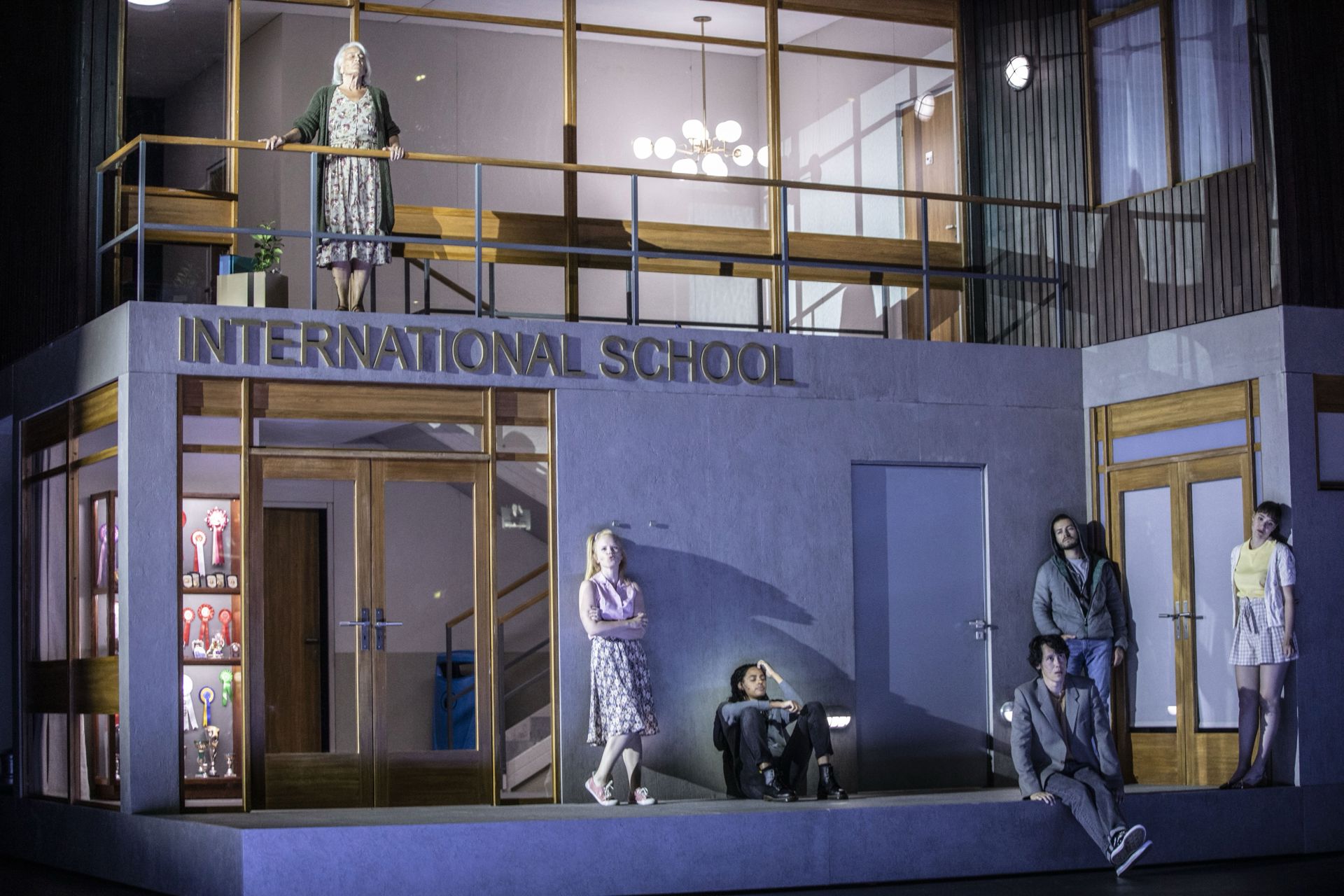 En teaterscene med en bygning som kulisse og skiltet “INTERNATIONAL SCHOOL” over indgangen. Flere personer står eller sidder spredt foran bygningen og på balkonen ovenover. De blålige scenelys og de stille, adskilte positioner giver scenen et køligt og afventende udtryk.