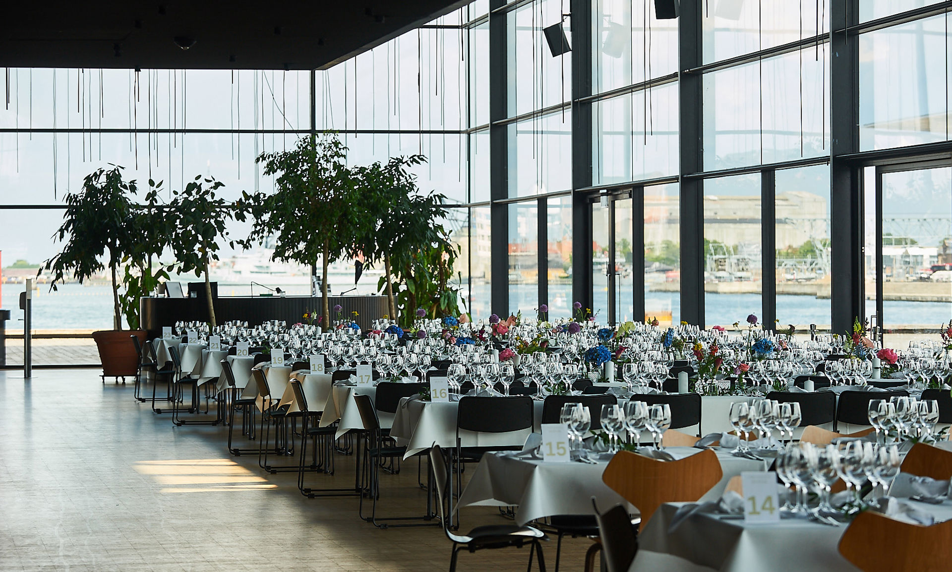 Skuespilhusets foyer dækket op til fest med lange borde, hvide duge, glas, service og lave blomsterdekorationer i flere farver. Store glasvægge giver udsigt til vand og bygninger udenfor. I den ene ende står store grønne planter i krukker, og rummet fremstår lyst, moderne og stilrent.