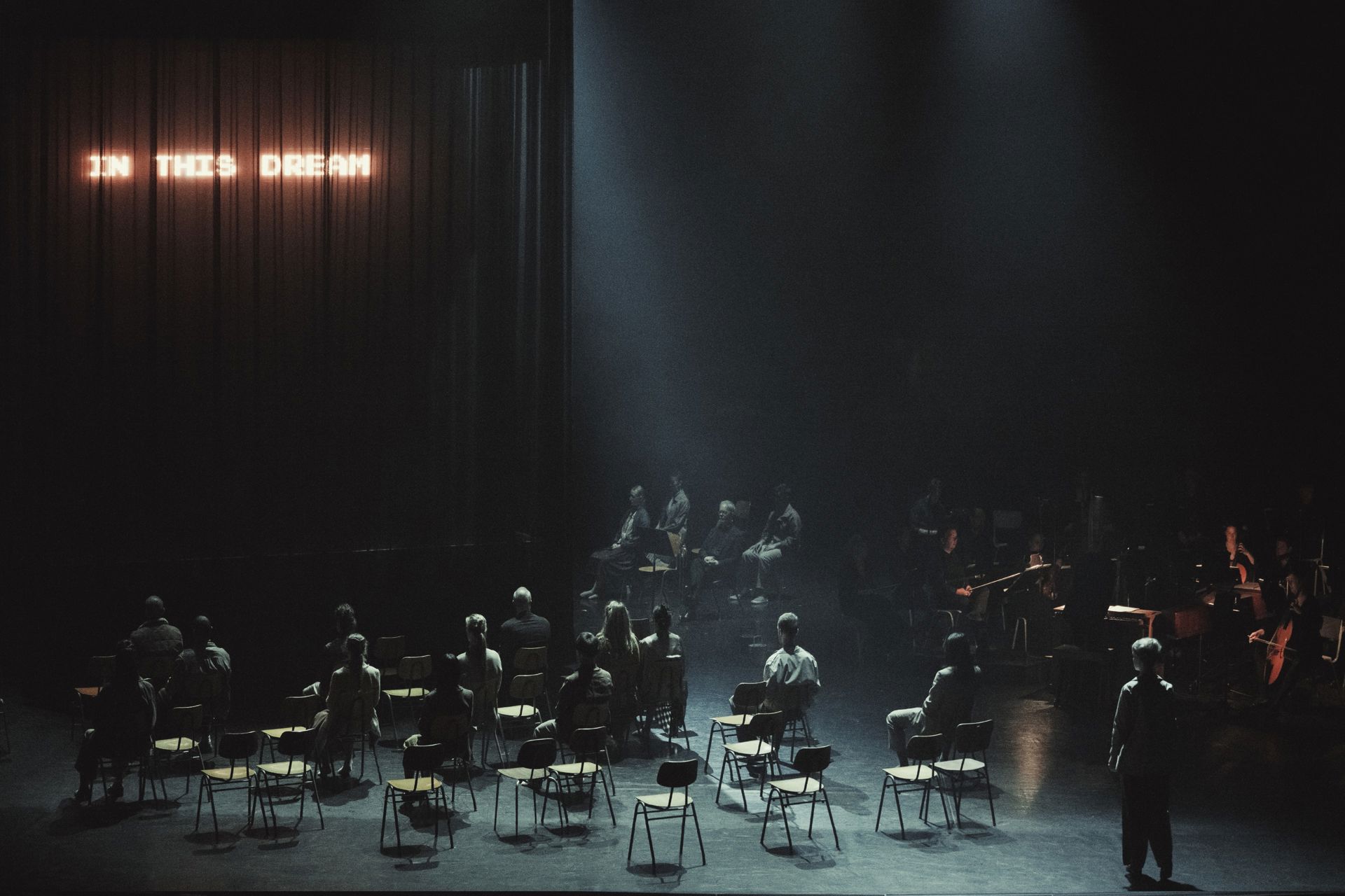 En mørk scene med mange stole og mennesker spredt rundt.  Forrest sidder og står personer enkeltvis med afstand. Midt på scenen er en tættere gruppe samlet i en svagt oplyst plet.  Bagtil anes flere personer ved instrumenter, og teksten “IN THIS DREAM” ses i lys på et mørkt tæppe.