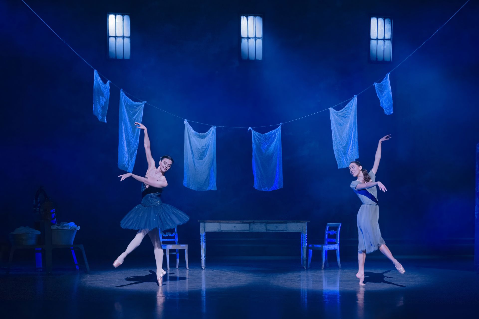 To balletdansere danser på en blåligt oplyst scene med vasketøj hængende på snore i baggrunden. Den ene danser til venstre i mørk nederdel med armen løftet, mens den anden til højre i lys kjole spejler bevægelsen. Rummet virker enkelt, roligt og poetisk.