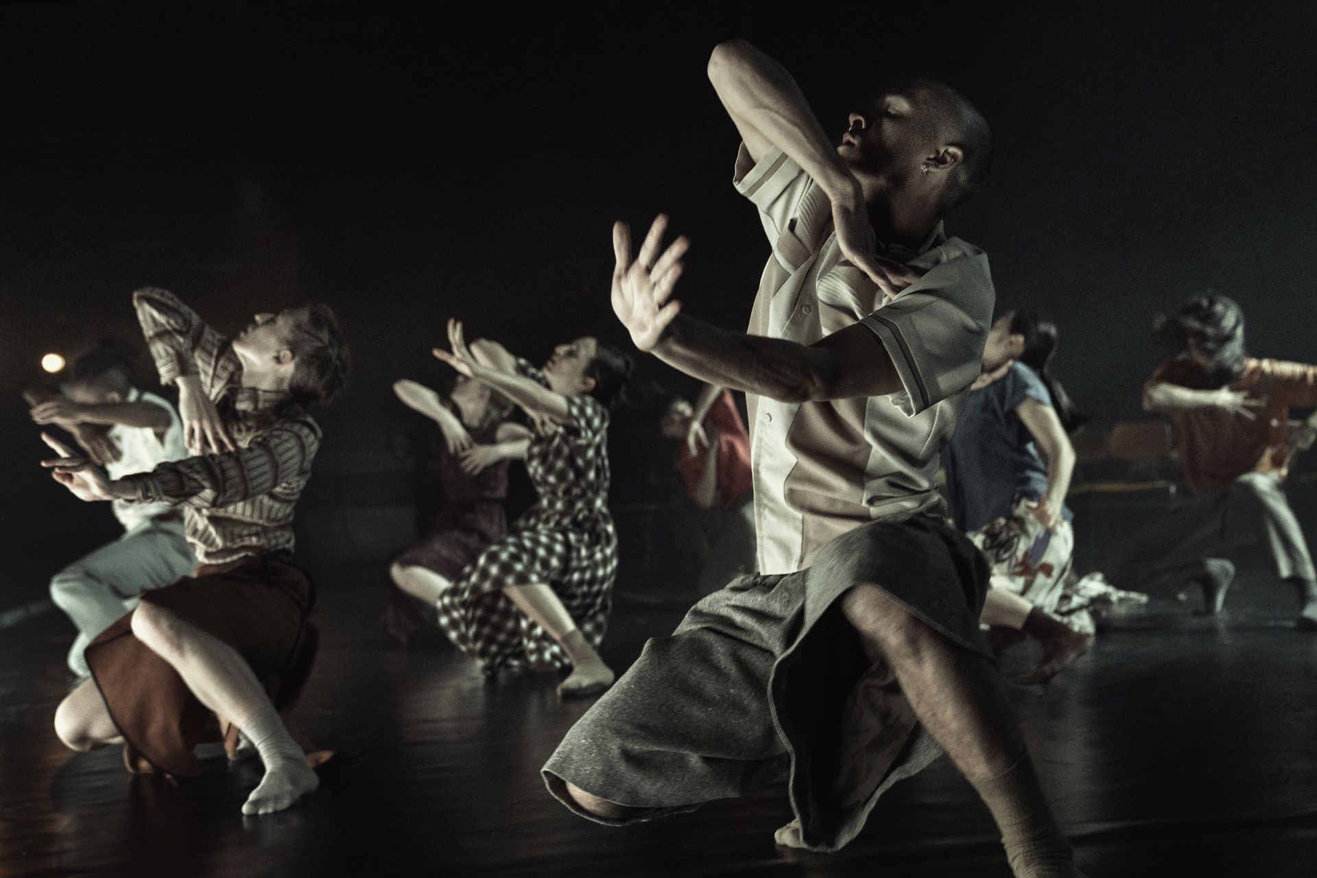 En gruppe dansere på en mørk scene midt i en intens koreografi.  Flere er i dramatiske, kantede bevægelser med arme og hænder løftet foran ansigt og over hovedet.  En forrest er bøjet dybt i knæene med brystet hævet og hovedet vendt op.  Bagved ses andre i forskellige stillinger med strakte fingre og spredte arme.  Lyset er svagt og koncentreret omkring gruppen, så baggrunden næsten forsvinder.
