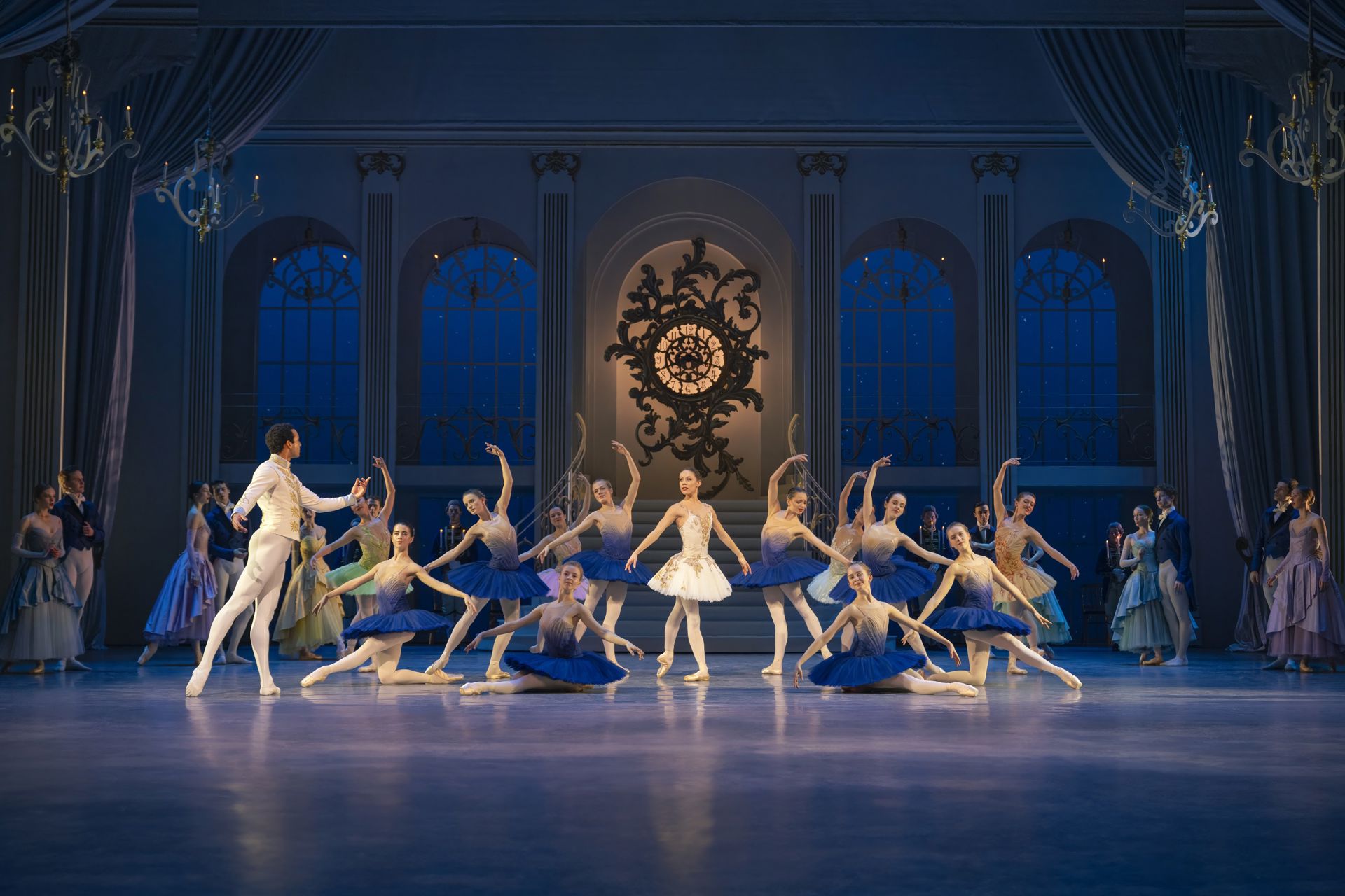 En balletscene i blåt lys med dansere samlet i en symmetrisk formation. Midt på scenen står en danser i hvidt, omgivet af dansere i blå og hvide kostumer, nogle knælende og andre med armene løftet. Bagved ses en klassisk balsal med ur og høje vinduer.