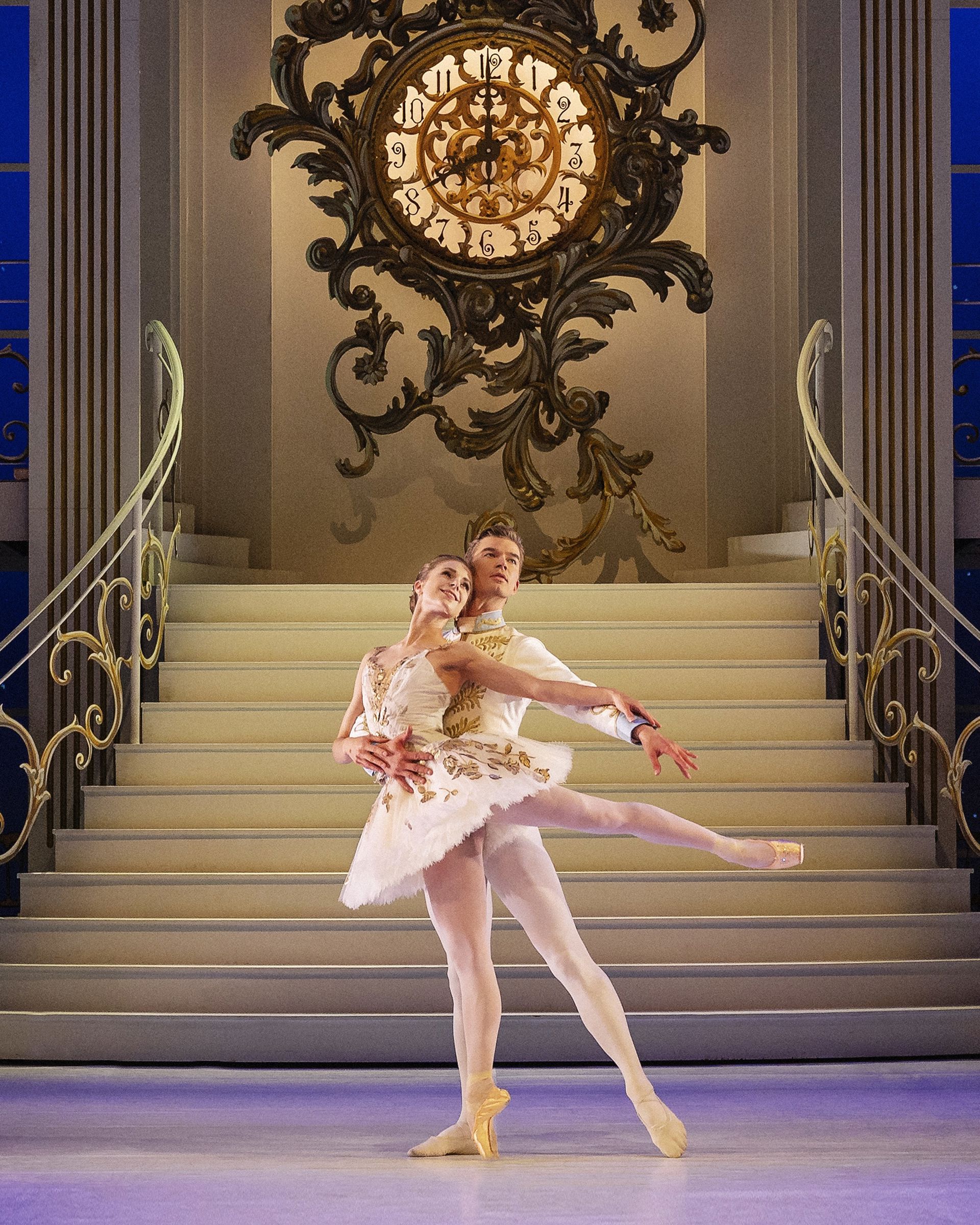 En balletdanser i lys, kort tutu danser foran en stor trappe og et dekorativt ur. Danseren står på tåspids med det ene ben løftet til siden og armene blødt placeret. Det lyse kostume og den klassiske scenografi giver billedet et elegant balletudtryk.