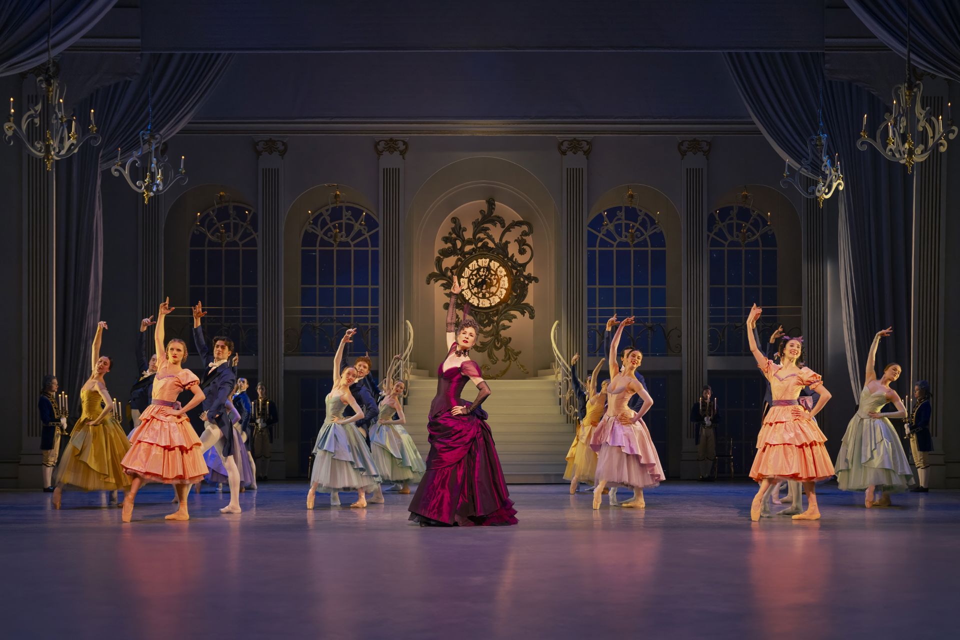 En balletscene i en stor balsal med dansere i pastelfarvede kostumer. I midten står en person i mørkerød kjole, mens dansere omkring løfter armene i elegante balletpositurer. Baggrunden har blå vinduer, trapper og et stort dekorativt ur.