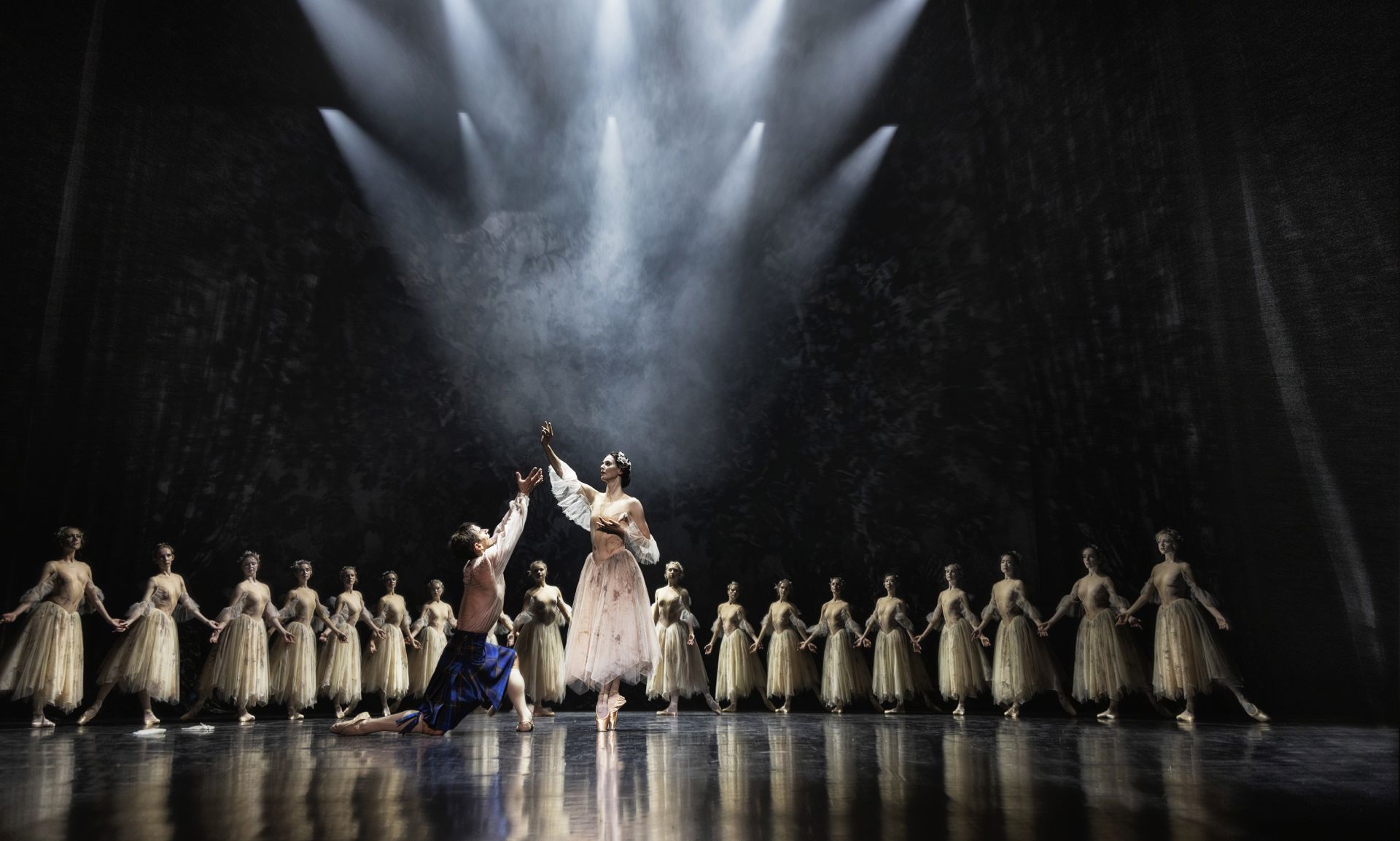 En balletscene med mange dansere under dramatisk scenelys. Forrest står to hoveddansere midt på scenen: én i mørkeblåt kostume knæler med en arm strakt op, mens en anden i en lys, let kjole står foran og rækker en arm op mod lyset. Bag dem danner en række dansere i lyse, ankellange kostumer en halvcirkel med hænderne forbundet. Lyset falder i koncentrerede kegler og fremhæver midten, mens baggrunden er mørk.
