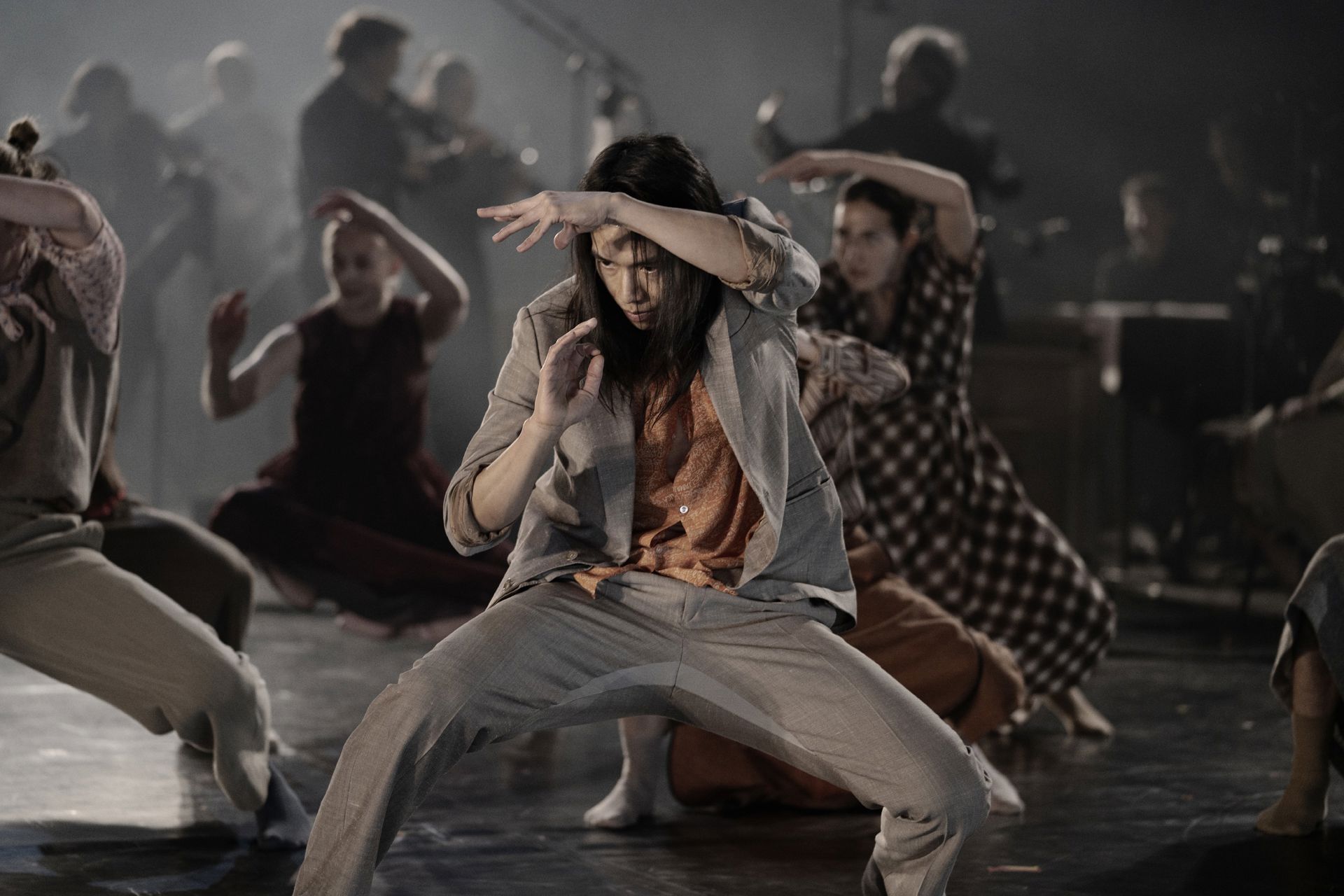 En moderne danseforestilling på en scene.  En danser i jakkesæt bøjer kroppen i en bred, koncentreret stilling med armene løftet.  Flere dansere bevæger sig omkring, og i baggrunden ses musikere i dæmpet lys.