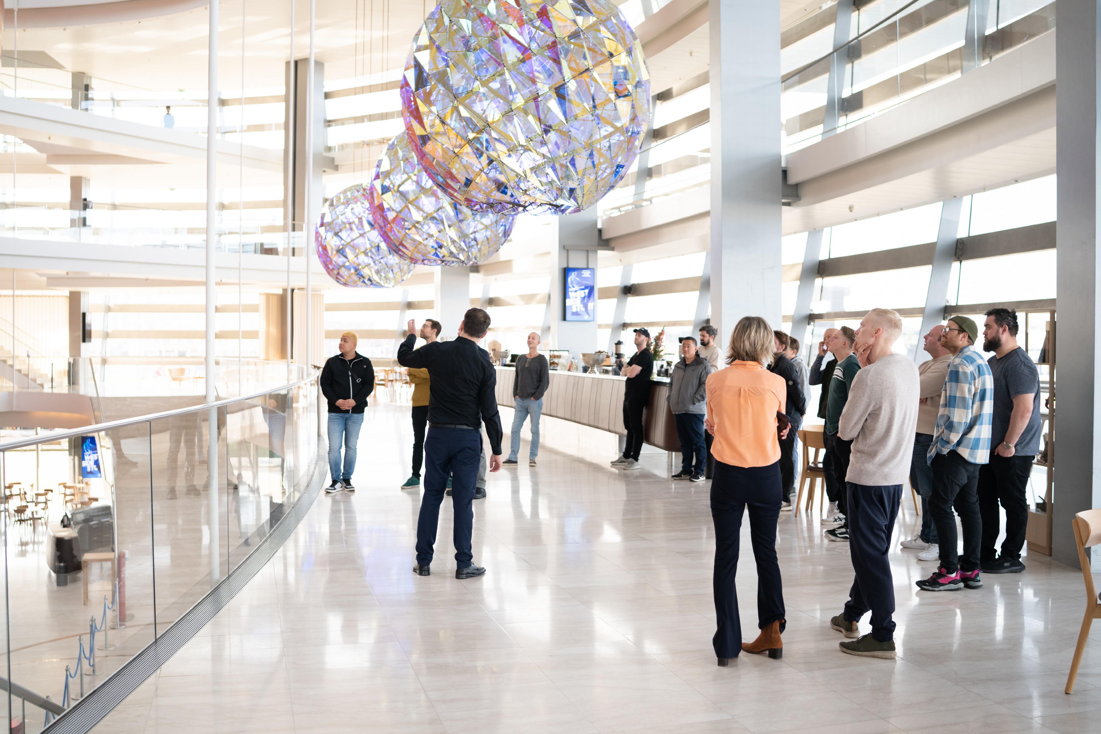 En lys og moderne indendørs foyer med en gruppe mennesker samlet på et blankt, lyst marmorgulv. I midten står en person i mørkt tøj og taler til gruppen foran sig, som står i en løs halvcirkel og lytter. I loftet hænger store, farverige, kugleformede installationer. Til højre ses en lang bar med enkelte personer bagved. Langs venstre side løber et glasrækværk, og i baggrunden ses flere åbne etager med meget dagslys.