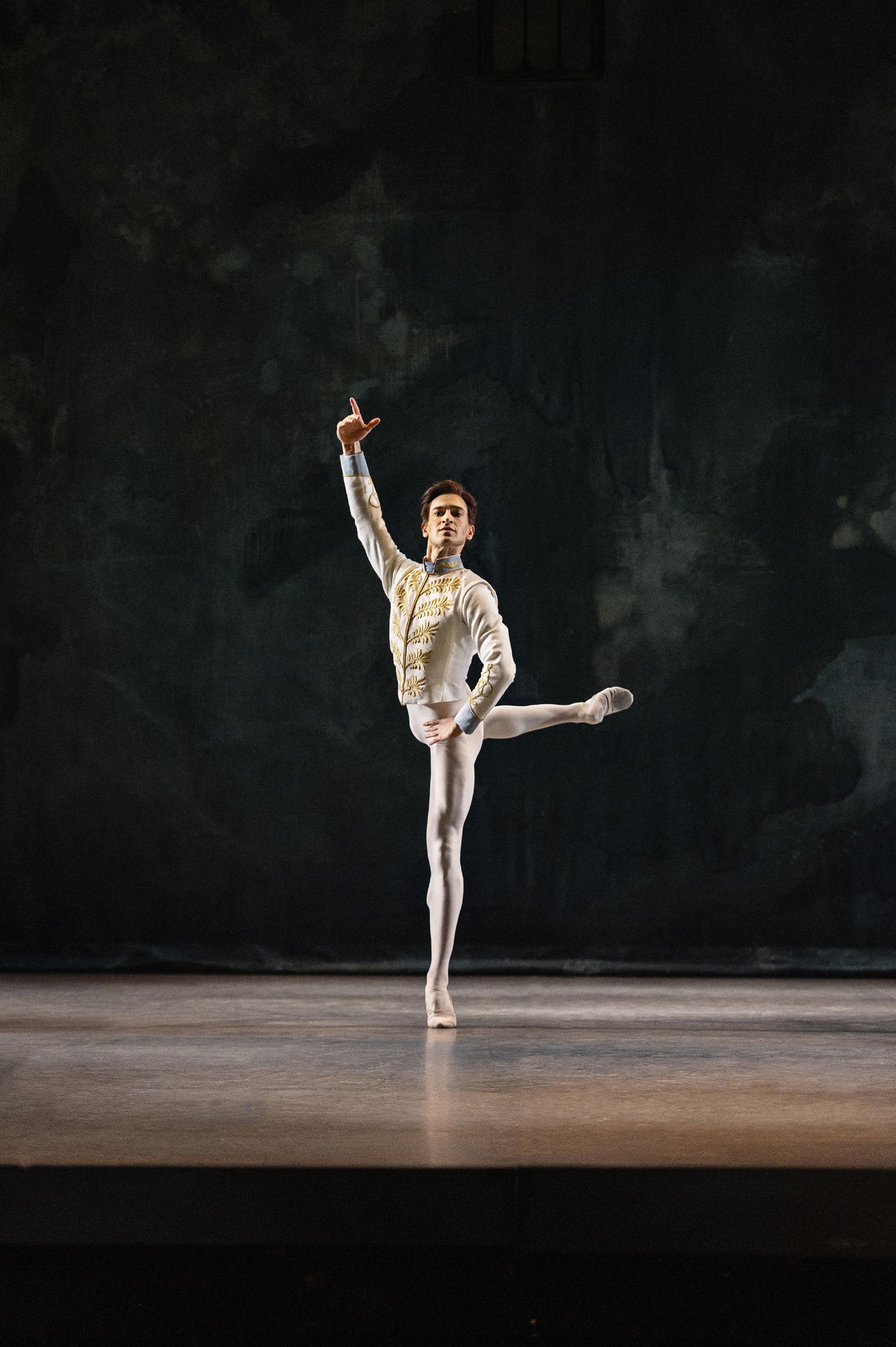 En balletdanser står alene på en mørk scene i en klassisk balanceposition. Det ene ben er løftet højt til siden, den ene arm peger op, og kroppen er strakt lodret. Det enkle lys og den mørke baggrund fremhæver danserens silhuet.