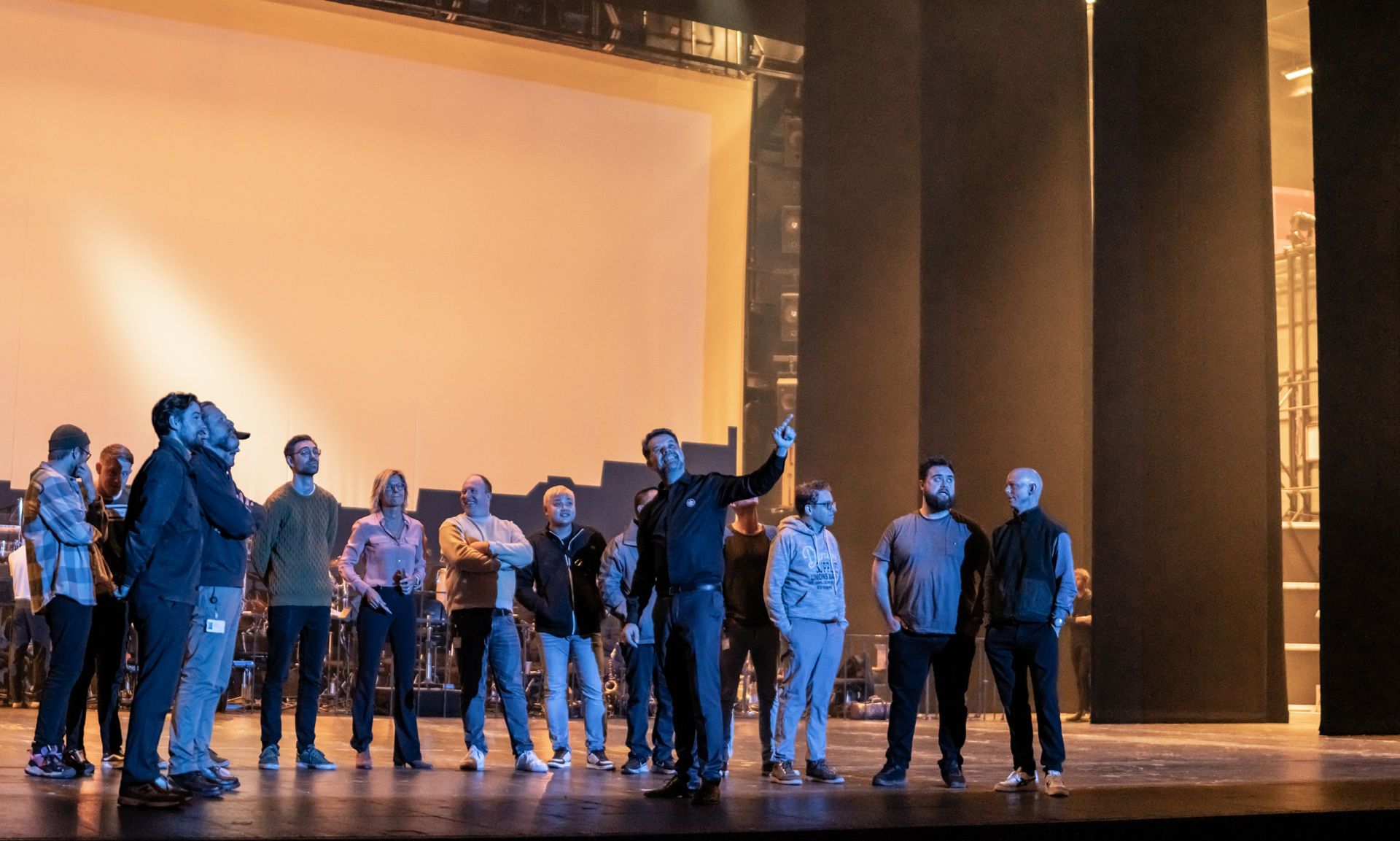 En gruppe mennesker står på en scene i Operaen. De er klædt i almindeligt hverdagstøj og står samlet, nogle tæt sammen og andre lidt for sig selv. Midt i gruppen står en person med armen løftet, som om noget markeres. Bag dem ses en stor, lyst oplyst bagvæg, mens scenegulvet foran er mørkere. I baggrunden anes udstyr eller instrumenter, som giver indtryk af en prøve eller forberedelse.