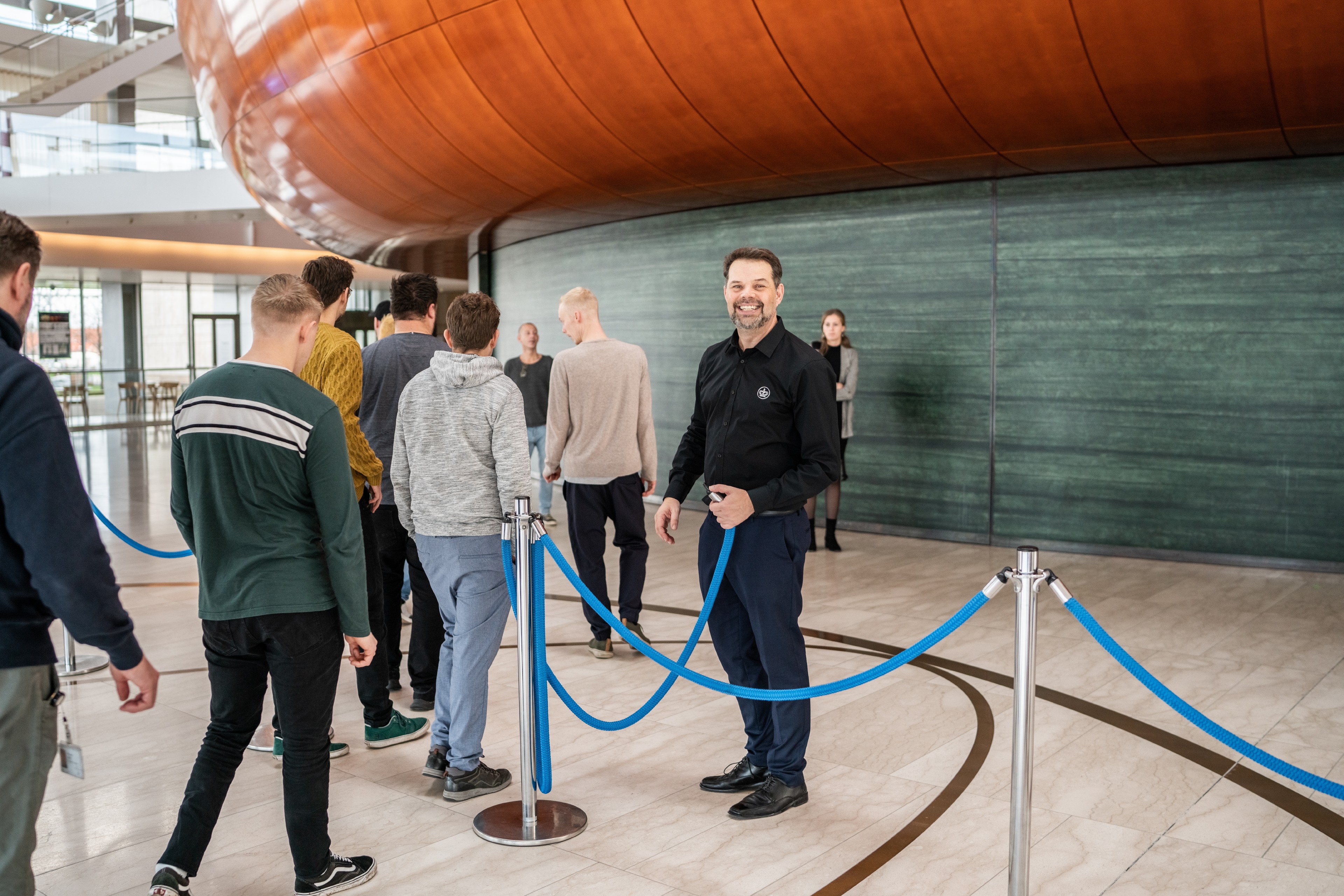 En gruppe personer står i en kø markeret med blå reb-afspærringer indendørs. Forrest til højre står en person i mørkt tøj og smiler mod kameraet, mens der holdes ved et af rebene. Foran står flere personer i afslappet tøj, vendt i samme retning, som om de venter på adgang. Gulvet er lyst og blankt, og rebene hænger i buer mellem sølvfarvede standere. I baggrunden ses en stor glat væg og en buet overflade øverst. Stemningen virker rolig og organiseret.